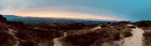 A panoramic view of rolling hills and valleys at sunset, with a winding dirt path leading through the landscape.