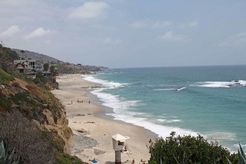 A scenic beach view with gentle waves, sandy shore, and distant cliffs under a partly cloudy sky.