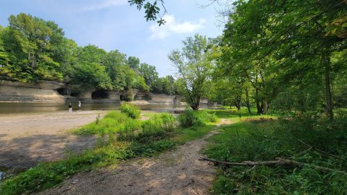 A serene riverside scene with lush greenery, rocky cliffs, and two people walking along a sandy path.