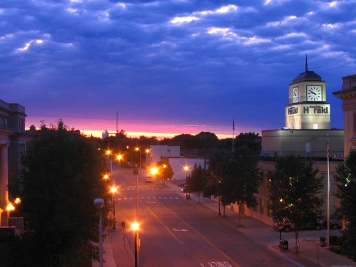 A city street at dusk, with a clock tower and streetlights illuminating the scene against a colorful sky.