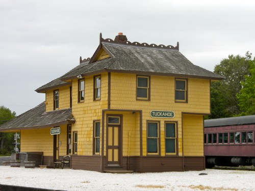 Historic yellow train station with "Tuckahoe" signage, surrounded by gravel and a vintage train car nearby.