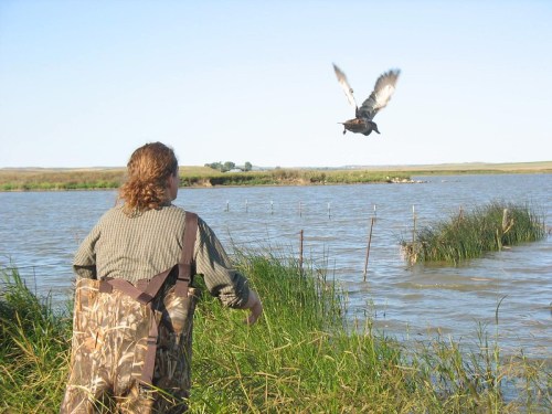 A person in camouflage waders watches a bird take flight over a calm lake surrounded by tall grass.