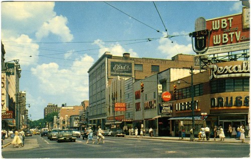 A busy city street scene with vintage cars, shops, and pedestrians under a blue sky with clouds.