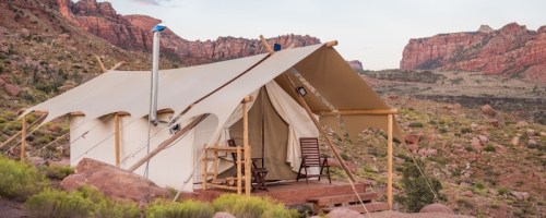 A canvas tent with a wooden deck and chairs, set against a backdrop of rocky mountains and desert landscape.