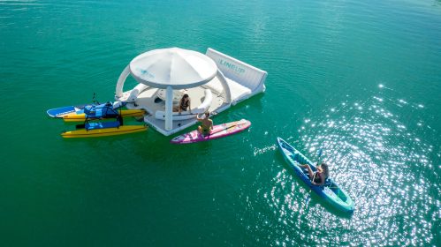 A floating platform in clear water with people on paddleboards and kayaks enjoying a sunny day.