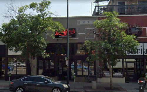 A street view of a restaurant with a neon sign, surrounded by trees and other storefronts.