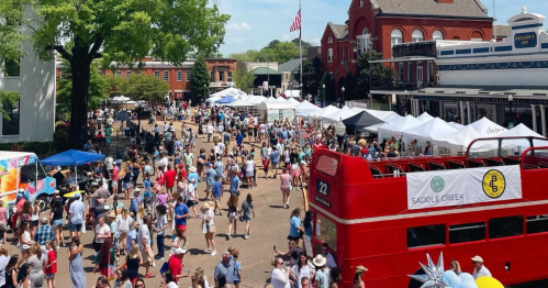 A bustling outdoor market scene with a crowd, tents, and a red double-decker bus in a lively town square.
