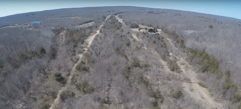 Aerial view of a winding dirt road through a wooded area, with sparse trees and open fields in the distance.