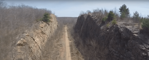 A dirt path runs between two rocky cliffs in a barren landscape, with sparse trees and a clear sky above.