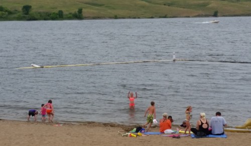A sandy beach with people relaxing and children playing in the water, with a boat in the background on a lake.