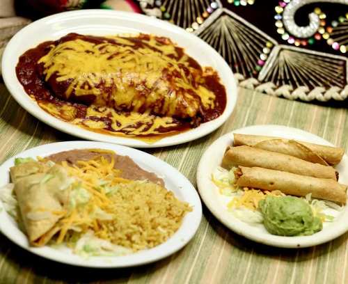 A plate of enchiladas topped with cheese and sauce, alongside taquitos, rice, and refried beans on a colorful tablecloth.
