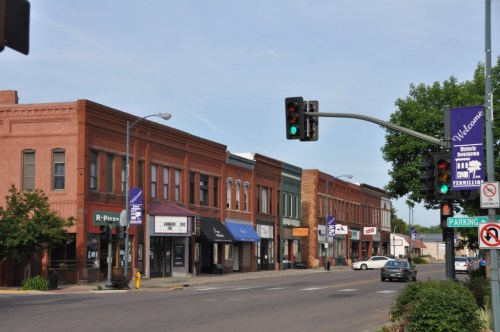 A street view of a small town with brick buildings, shops, and traffic lights under a clear blue sky.
