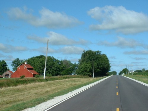 A rural road stretches ahead, flanked by green fields and a red barn under a blue sky with fluffy clouds.