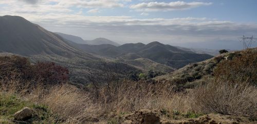 A scenic view of rolling hills and valleys under a cloudy sky, with sparse vegetation and distant mountains.