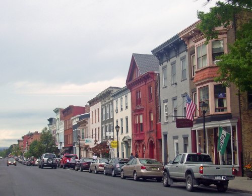 A charming street lined with historic buildings, shops, and parked cars under a cloudy sky.