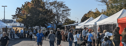 A bustling outdoor market with people browsing tents and stalls under clear blue skies and autumn trees.