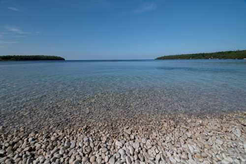 A serene shoreline with smooth pebbles leading to calm, clear water under a blue sky. Green hills in the distance.