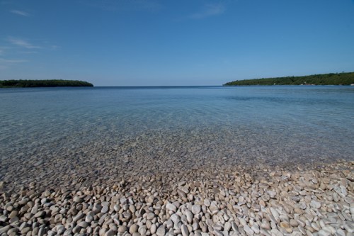 A serene shoreline with smooth pebbles leading to calm, clear water under a blue sky. Green hills in the distance.