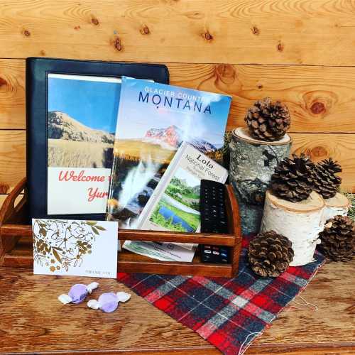 A wooden tray holds a travel guide to Montana, a remote, a card, and pinecones on a plaid fabric against a wooden backdrop.