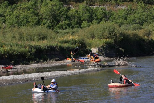 A group of people kayaking and canoeing on a river, surrounded by lush greenery and a rocky shoreline.
