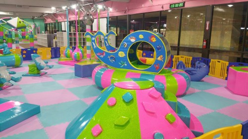 Colorful indoor play area with various climbing structures and soft play equipment in vibrant pink, green, and blue.