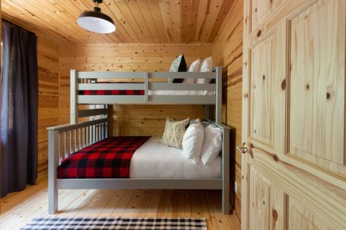 Cozy wooden room featuring a bunk bed with plaid bedding and a door, illuminated by a ceiling light.