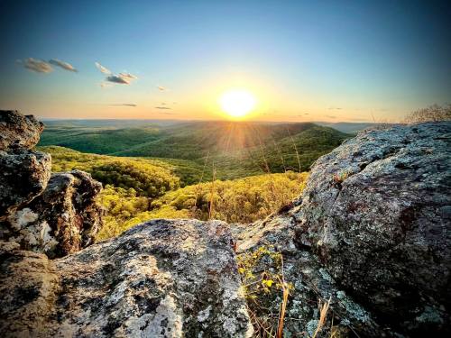 Sunset over a lush green valley, viewed from rocky terrain, with warm light illuminating the landscape.