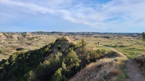 A scenic landscape with rolling hills, greenery, and a winding path, under a blue sky with wispy clouds.