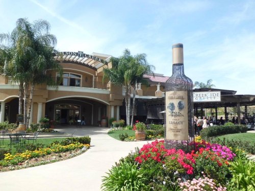 A winery entrance featuring a large wine bottle sculpture surrounded by colorful flowers and palm trees.