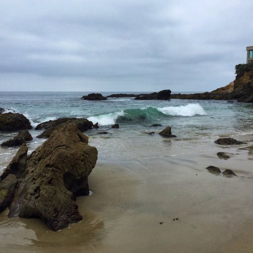 A rocky beach with gentle waves under a cloudy sky, featuring calm waters and distant cliffs.