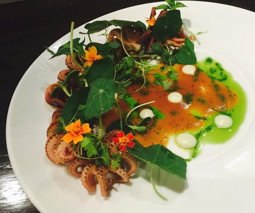 A gourmet dish featuring octopus, vibrant sauces, and edible flowers on a white plate, garnished with fresh greens.