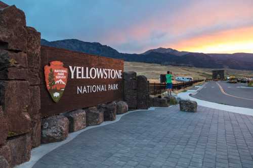 A person stands near the Yellowstone National Park entrance sign at sunset, with mountains in the background.