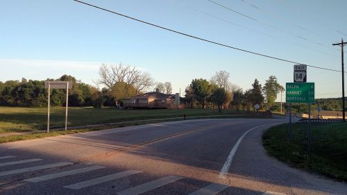 A rural road curves with a sign indicating distances to nearby towns and a house in the background under a clear sky.