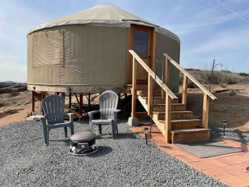 A cozy yurt with wooden steps, surrounded by gravel, featuring two chairs and a fire pit in a scenic outdoor setting.
