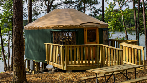 A wooden yurt with a tan roof, surrounded by trees, overlooking a lake, with a deck and seating area.