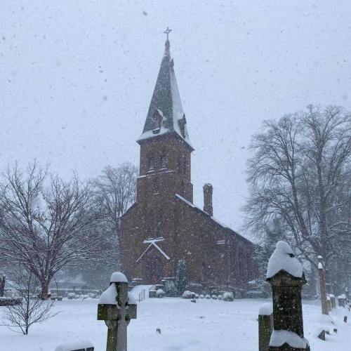 A snow-covered church with a tall steeple, surrounded by gravestones in a winter landscape. Snowflakes are falling heavily.