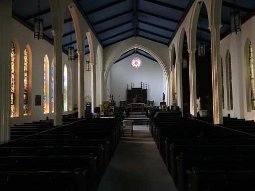 Interior of a church featuring stained glass windows, wooden pews, and a simple altar at the front.