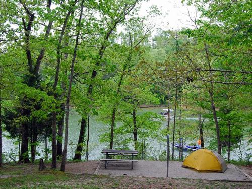 A serene campsite by a lake, featuring a yellow tent, picnic table, and lush green trees surrounding the water.