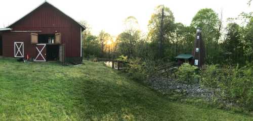 A red barn at sunset, surrounded by greenery and a small pond, with a chimney and trees in the background.