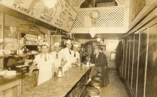 Historic diner interior with five men in white aprons, a long counter, and vintage decor.