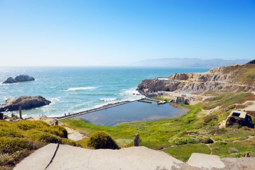 A scenic coastal view featuring a calm ocean, rocky cliffs, and a small pond surrounded by greenery under a clear blue sky.