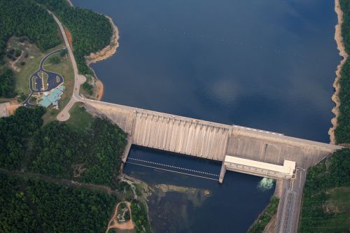 Aerial view of a large dam with water reservoir, surrounded by green trees and a small building nearby.