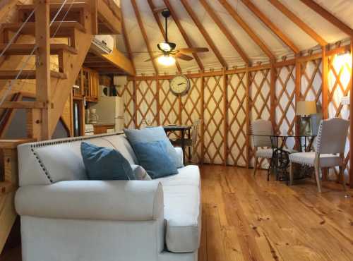 Cozy interior of a yurt featuring a sofa, dining table, and wooden accents, with a staircase and ceiling fan.