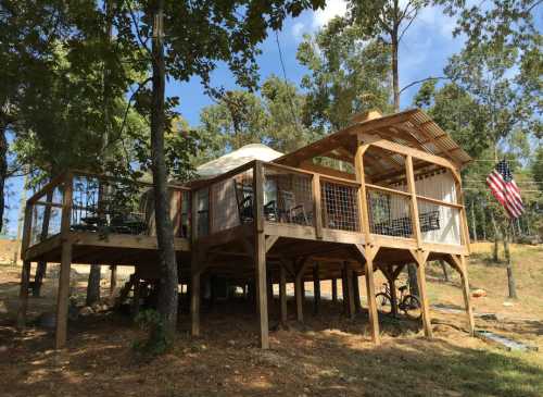 A wooden cabin on stilts surrounded by trees, featuring a deck and an American flag.