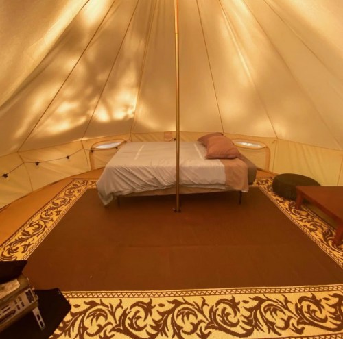 Cozy interior of a glamping tent featuring a bed, rug, and wooden furniture. Warm lighting creates a welcoming atmosphere.