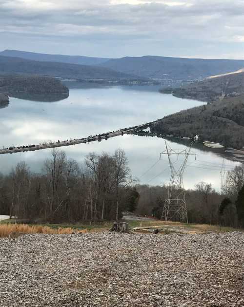 Scenic view of a lake surrounded by mountains, with a power line tower in the foreground and cloudy skies above.