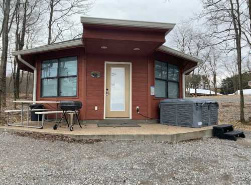 A modern cabin with large windows, a grill, and a hot tub, surrounded by trees and gravel.