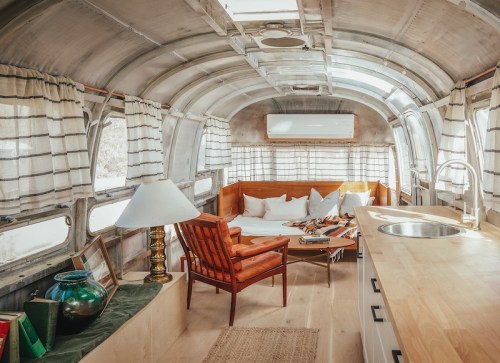 Cozy interior of a vintage Airstream trailer with a wooden sofa, lamp, and kitchen area, featuring large windows.