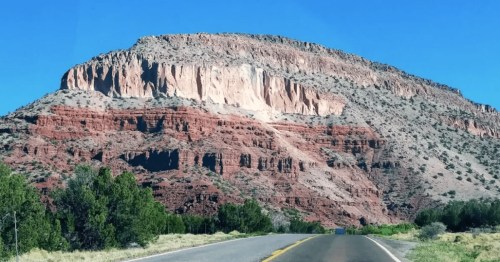 A scenic view of a rocky, red mountain under a clear blue sky, with a road leading towards it and green shrubs in the foreground.