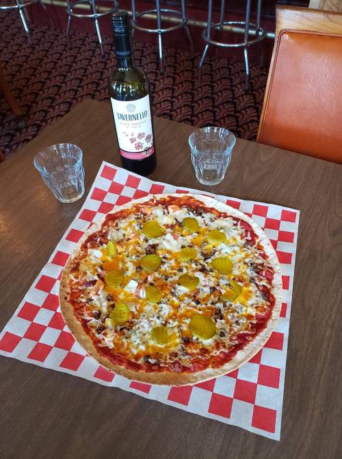 A large pizza topped with pickles, cheese, and other ingredients, accompanied by a bottle of wine and two glasses.